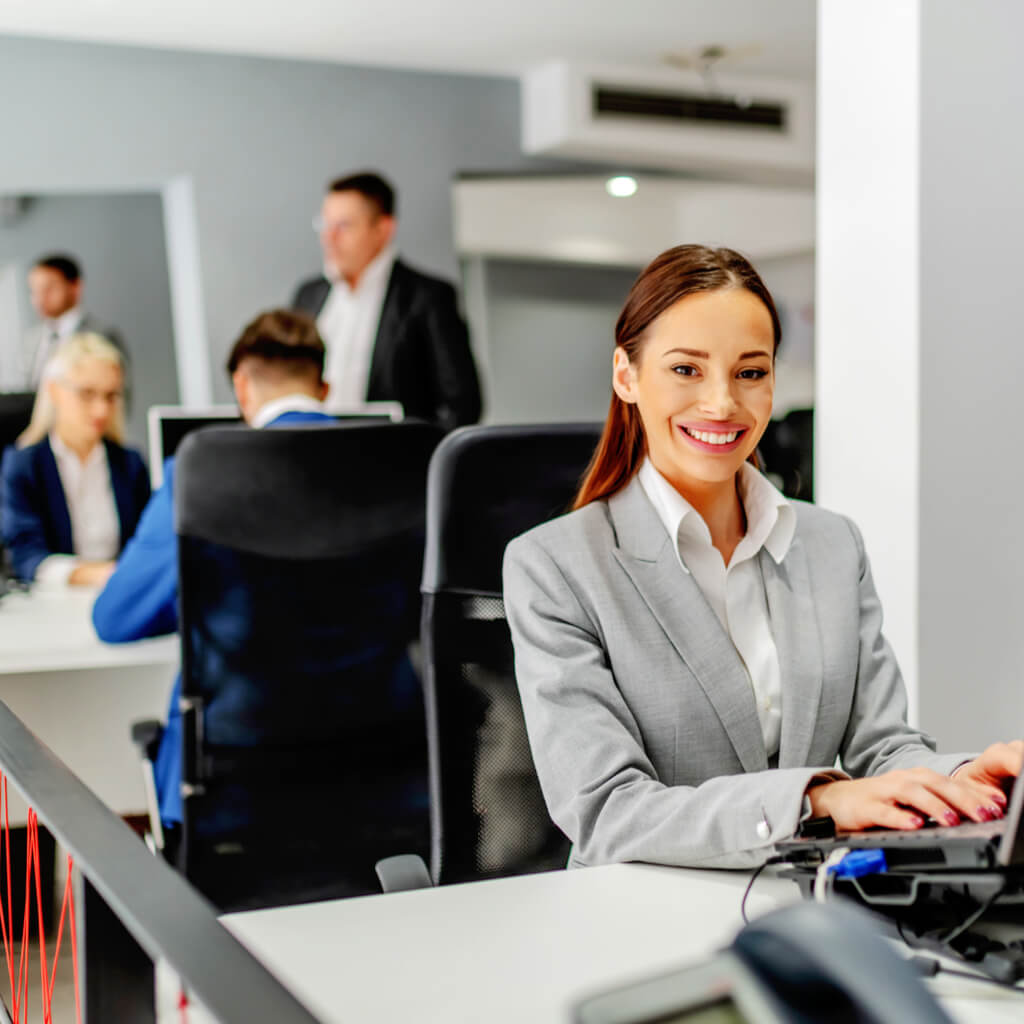 data entry outsourcing - Image Shutterstock-1735831754 - Young positive dedicated caucasian employee in suit sitting at her work place and using laptop for data entry. In background are her colleagues working.