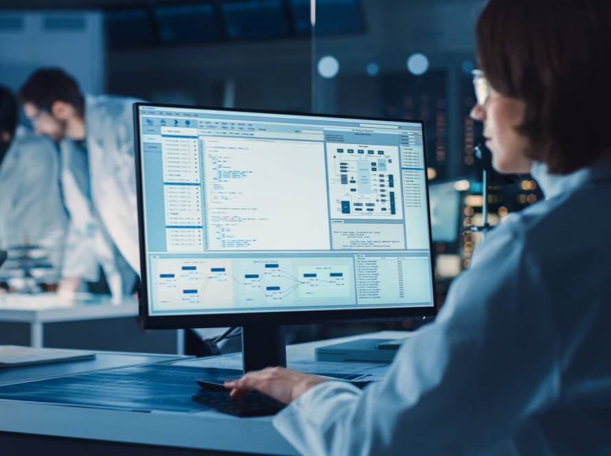 Billing and coding - featured image - Over the Shoulder Shot: Female IT Scientist Uses Computer Showing System Monitoring and Controlling Program. In the Background Technology Development Laboratory with Scientists, Engineers Working