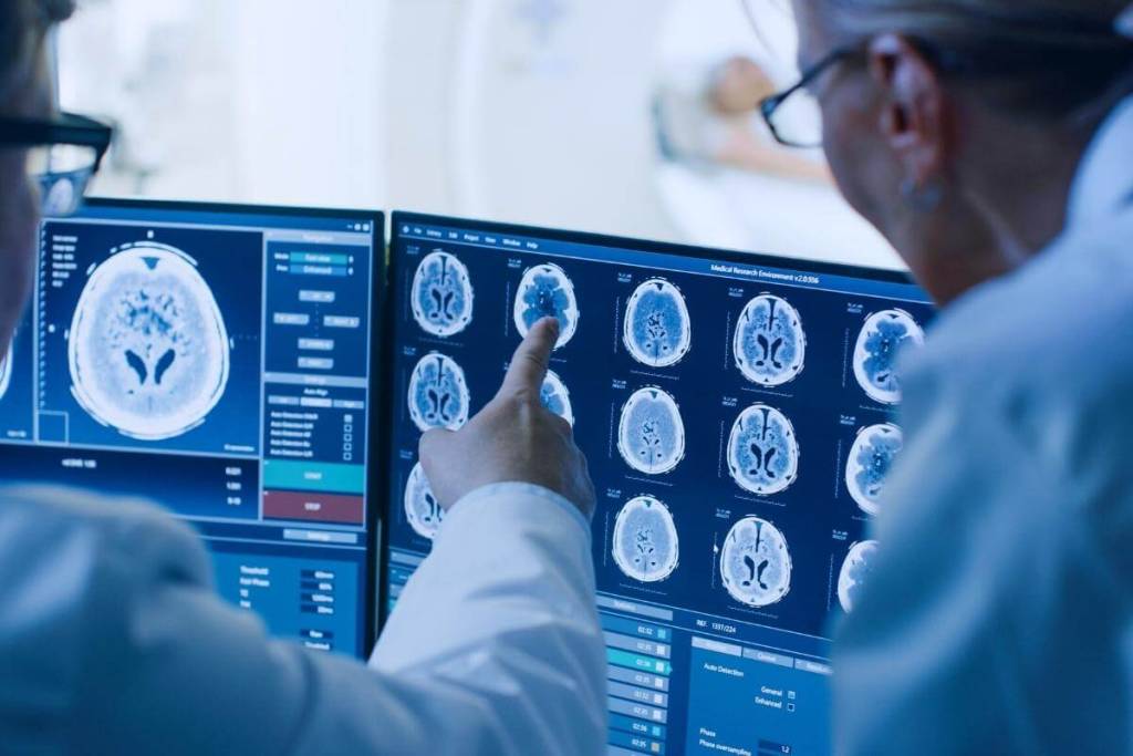 Oncology Medical Billing trends - In Control Room Doctor and Radiologist Discuss Diagnosis while Watching Procedure and Monitors Showing Brain Scans Results, In the Background Patient Undergoes MRI or CT Scan Procedure.