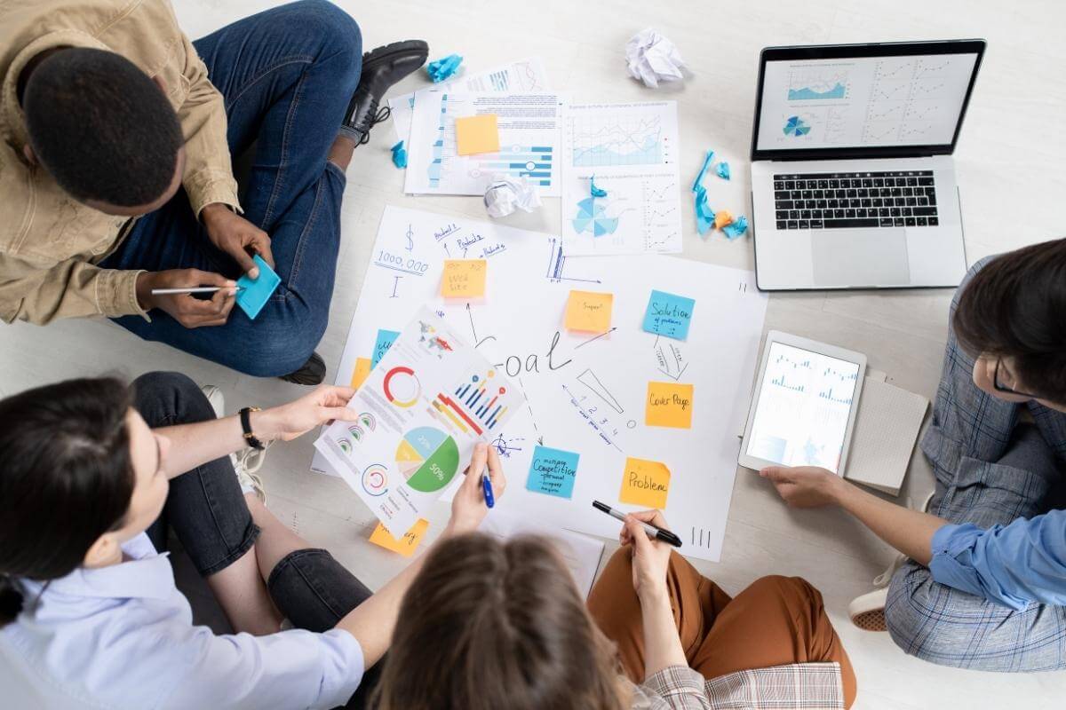 Above view of multi-ethnic collegues sitting on floor with papers and devices and discussing business plan.