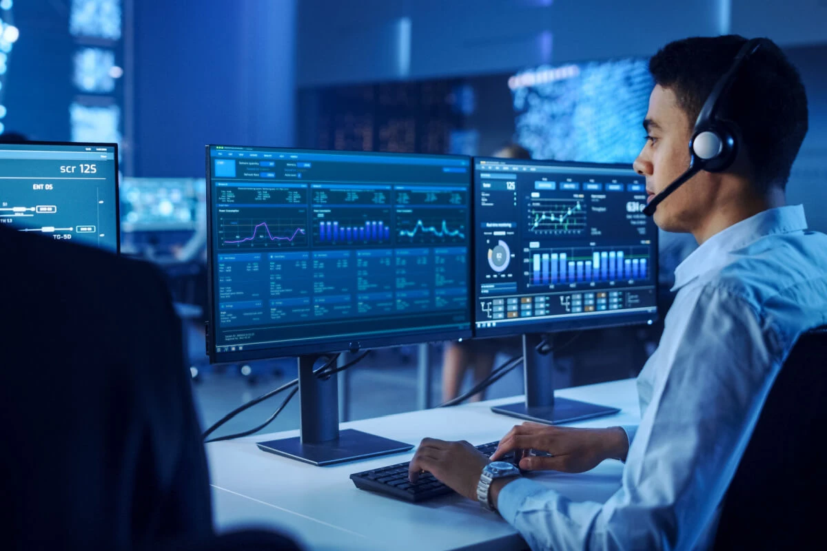 Confident Male Data Scientist Works on Personal Computer Wearing a Headset in Big Infrastructure Control and Monitoring Room. Young Engineer in a Business Call Center Office Room with Colleagues.