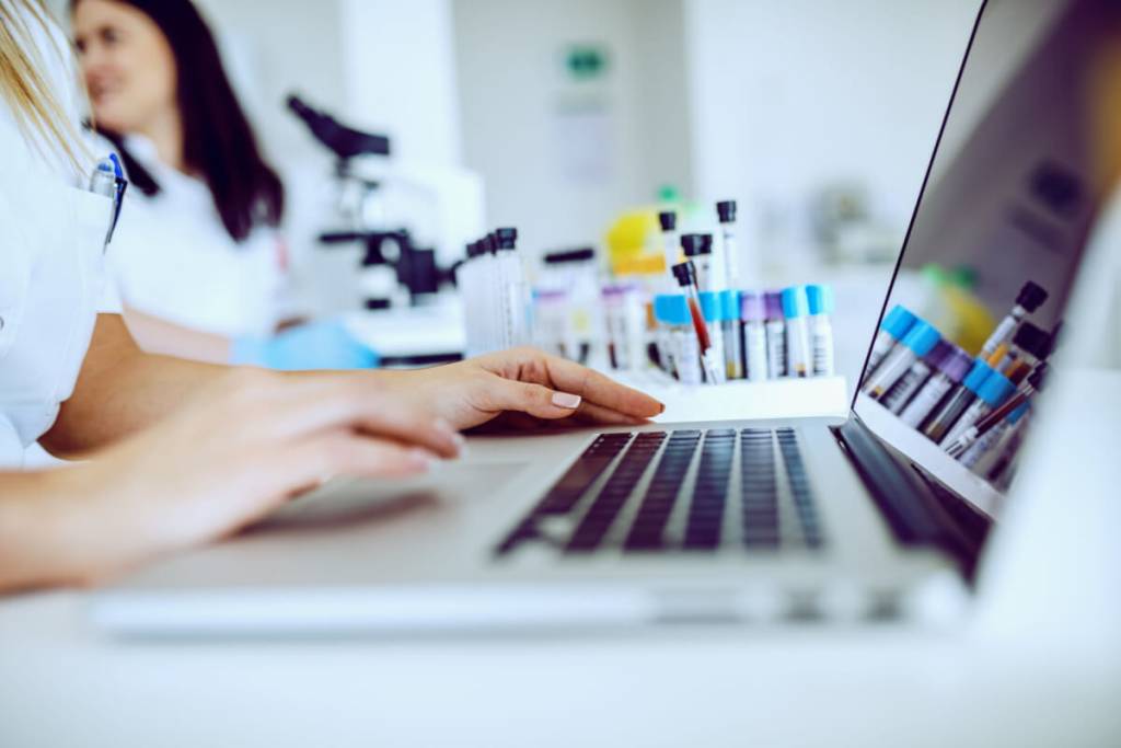 Medical Data Entry Services- Featured Image_Close up of female lab assistant in white uniform sitting in lab and using laptop for data entry. In background is her colleague working.