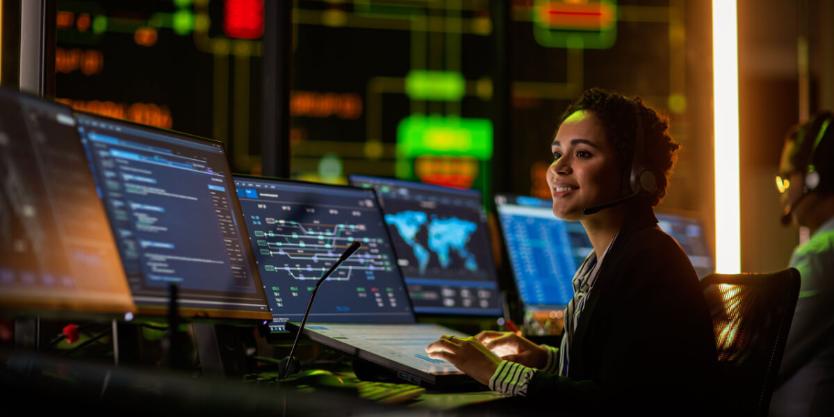 Multiethnic Female IT Technical Support Specialist Talking with a Client in Headphones and Working on Desktop Computer in Monitoring Control Room with Big Digital Screens with Server Blockchain Data.